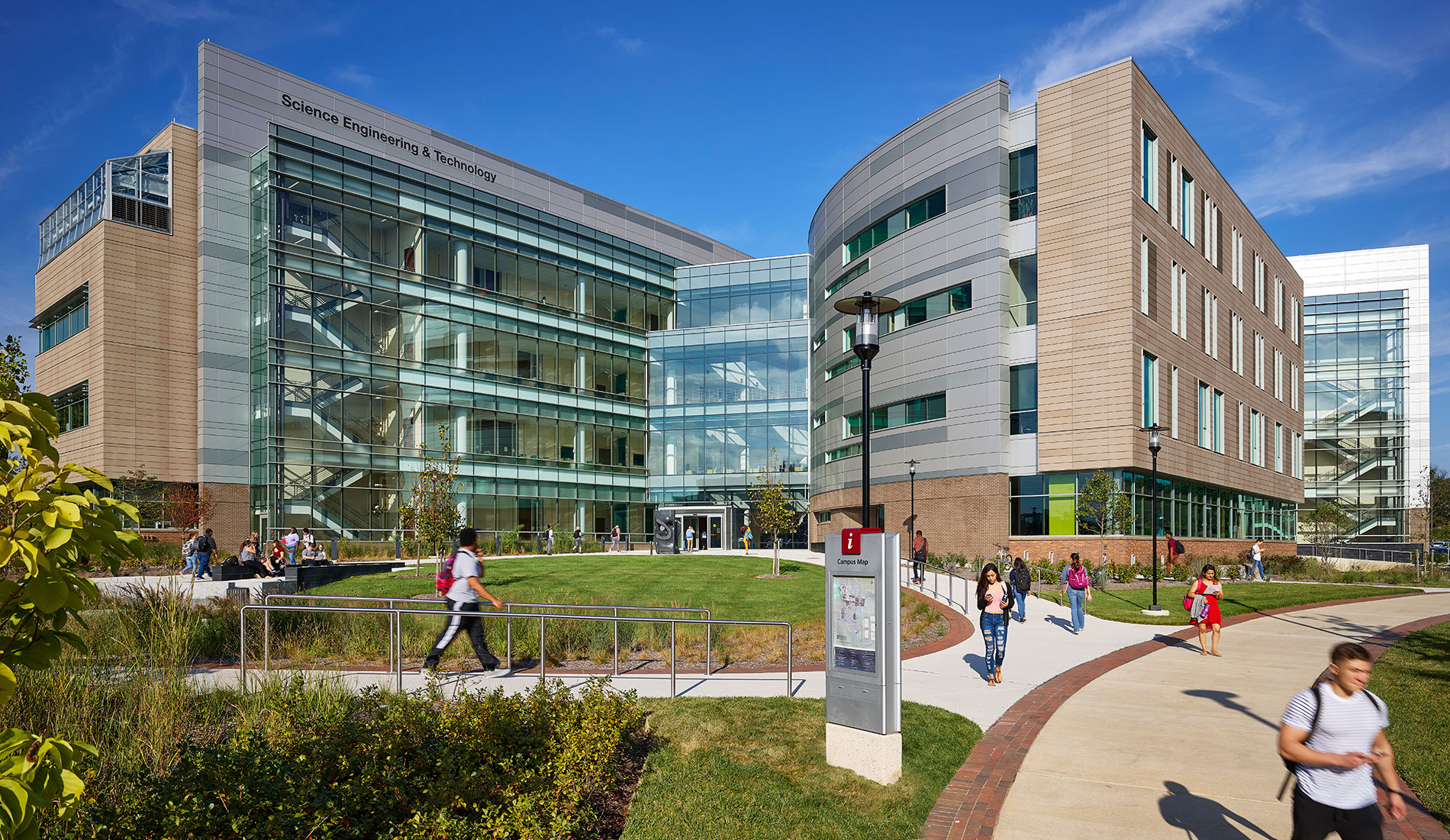 Exterior view of the Science, Engineering, and Technology Building at Howard Community College. The modern structure features large glass windows, brick and metal paneling, and multiple stories. Trees and landscaped greenery surround the entrance, and the sky is clear in the background.”