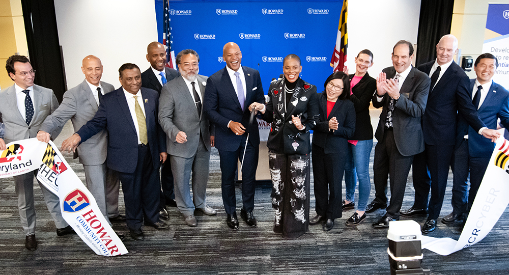 Group photo of Governor Moore, Daria Willis, government officials, business leaders, and educators at a ribbon-cutting ceremony for Howard Community College's new cybersecurity training center. The participants stand smiling in front of a blue backdrop with the college's logo, holding a ribbon with Maryland branding, signifying the event's official opening.