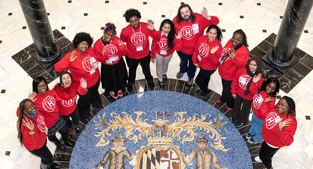 Group of Howard Community College students and staff wearing matching red shirts stand in a circle indoors and wave up at the camera.