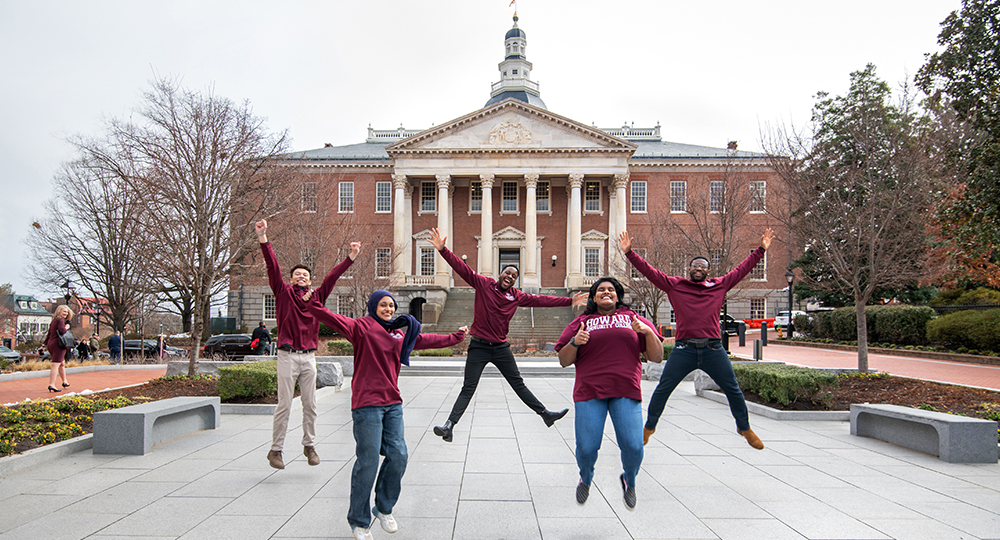 Five diverse students in maroon Howard Community College shirts jump and pose excitedly in front of a building with a dome in Annapolis, surrounded by walkways, benches, and trees.