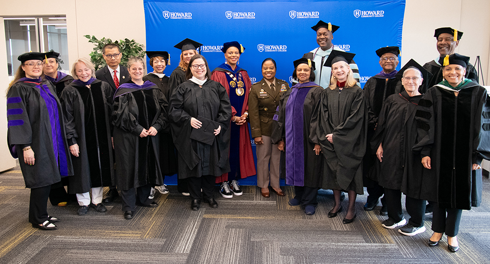 group of HCC staff and guests dressed in cap and gown standing in front of the blue step and repeat with HCC logos on the fabric