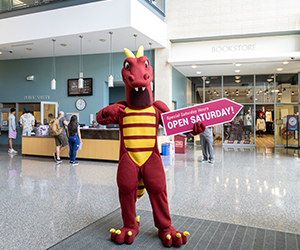 HCC's red dragon campus mascot holds a “Super Saturday – Open Saturday!” sign inside a college student center with the bookstore and students in the background.