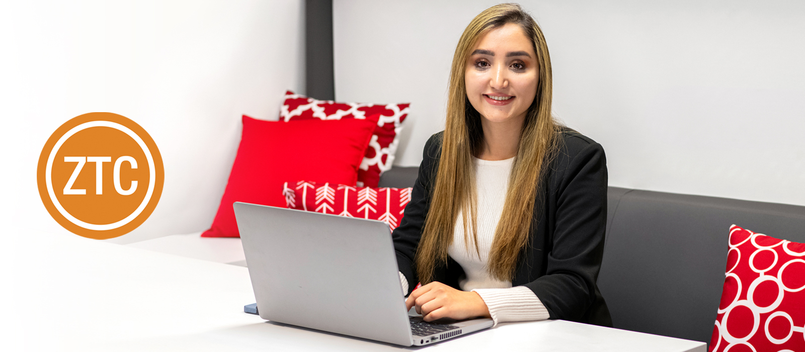 Smiling student with long hair wearing a black blazer and white top sits at a table with a laptop, in a modern lounge area with red and white pillows. An orange 'ZTC' logo is visible on the left side of the image.