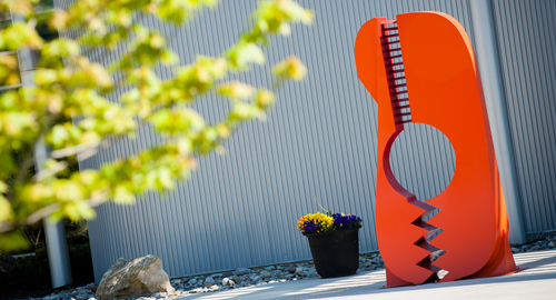 Bright orange-red abstract metal sculpture with a circular cutout and serrated lower edge and a small pot of yellow and purple flowers at its base, set against a corrugated metal wall
