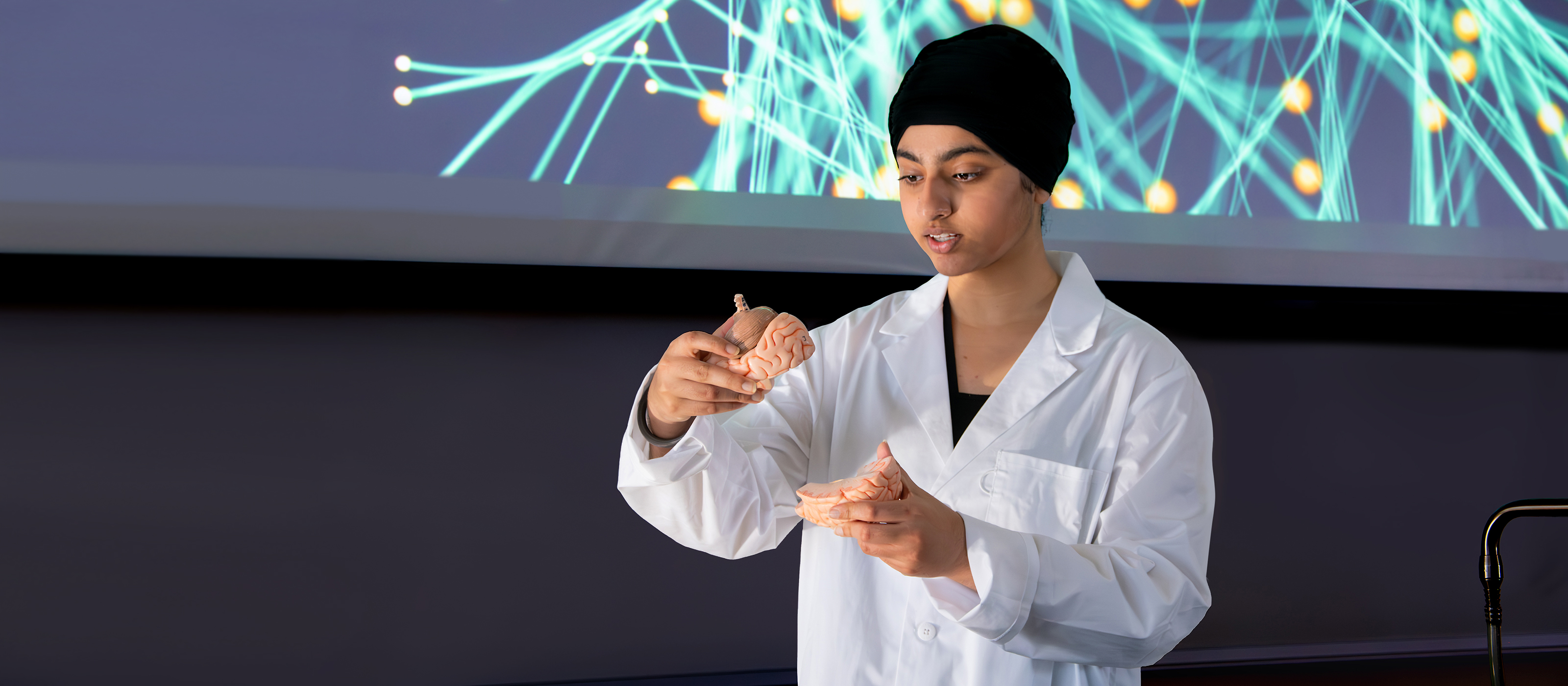 A person in a lab coat examines a brain model in front of a screen displaying neural connections. The scene conveys focus and scientific inquiry.