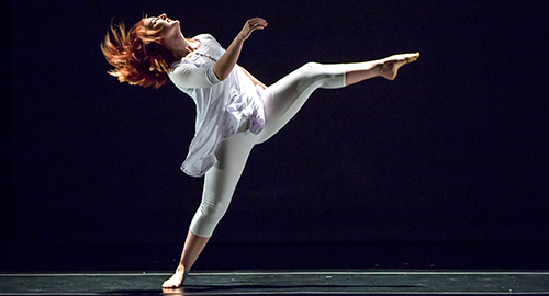 Dancer in a flowing white tunic balancing on one leg on a dark stage with arms raised in an expressive pose