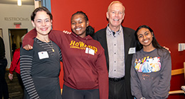 Four people standing close together and smiling at the camera inside a building. One young woman is wearing a Howard Community College sweatshirt, standing between two adults and another young woman in a colorful graphic sweatshirt at the Student Donor Luncheon..