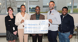 Five smiling individuals standing indoors, holding a large ceremonial check made out to 