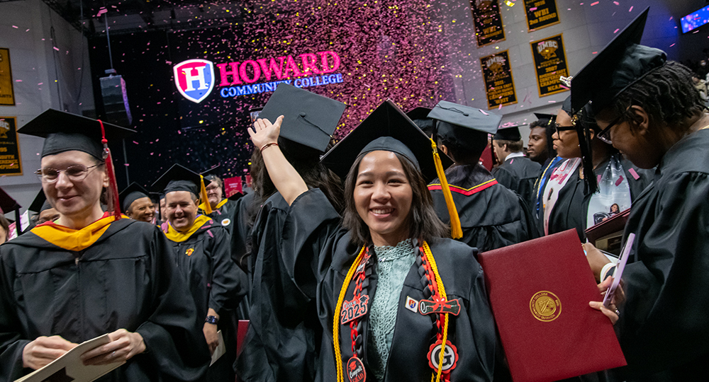 Graduates celebrate at Howard Community College's commencement ceremony. A smiling graduate in the foreground raises her arm in excitement, wearing a decorated graduation cap, honor cords, and holding a diploma folder. Confetti fills the air, and a large Howard Community College sign is visible in the background.