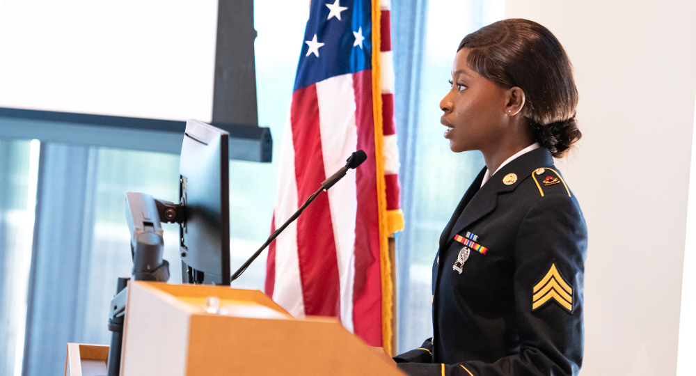 A U.S. Army servicewoman in dress uniform speaks at a podium with a microphone. She is standing beside a U.S. flag, and a computer monitor is in front of her. The setting is the Military and Honors Cording event.