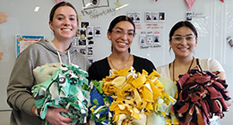 Three young women standing in a classroom, smiling and holding colorful handmade fleece blankets. The background features a whiteboard with photos and decorations.
