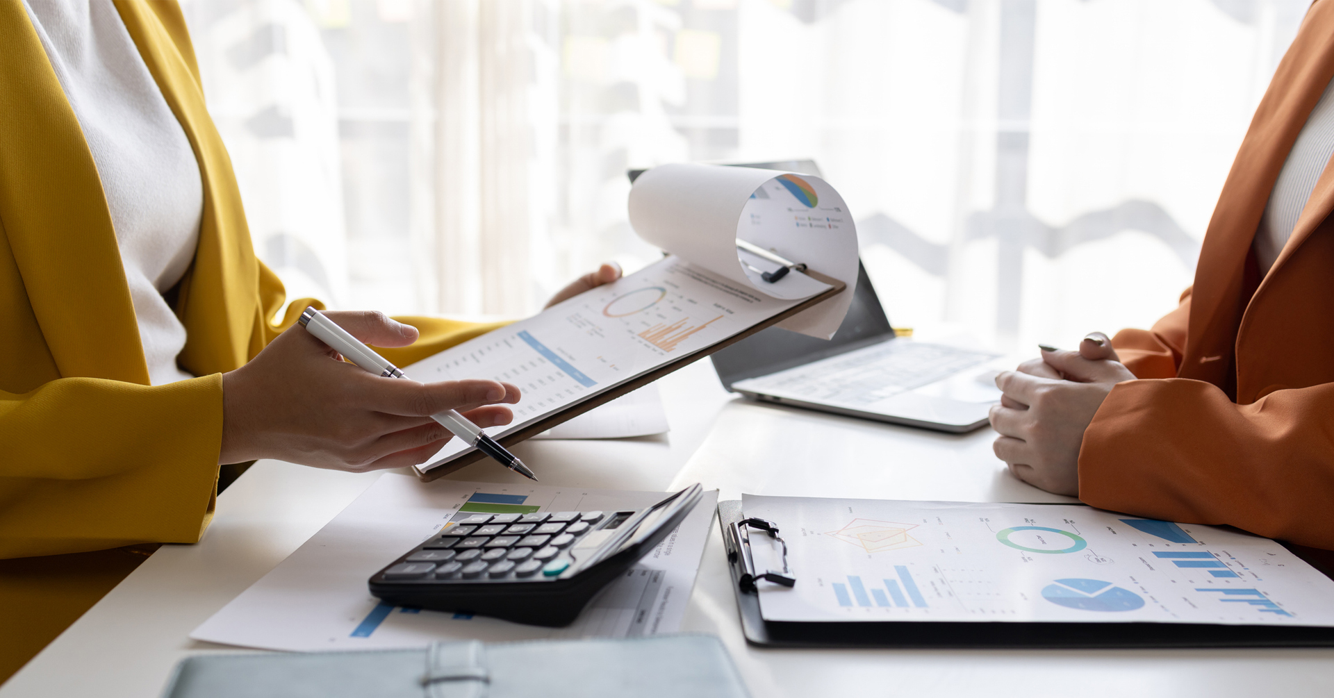 Two professionals in business attire, one wearing a yellow blazer and the other an orange blazer, are engaged in a discussion at a desk. One person is holding a clipboard with financial charts and graphs, while the other listens attentively. A calculator, additional documents with graphs, and a laptop are also on the table, indicating a financial or business analysis setting.