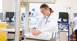 A healthcare professional in a white coat practices infant care on a mannequin baby in a clinical lab setting, surrounded by medical equipment and computer monitors.