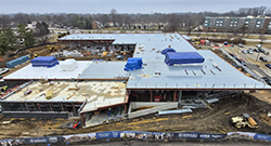 Aerial view of a large building under construction on a college campus, showing a wide roof with several blue-covered sections, exposed framing on one side, and construction equipment and materials surrounding the site.