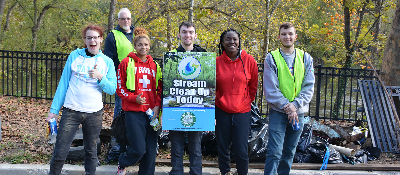 Group of volunteers smiling together after a successful stream cleanup. They stand outdoors with a sign reading 'Stream Clean Up Today,' surrounded by filled trash bags and autumn trees in the background.