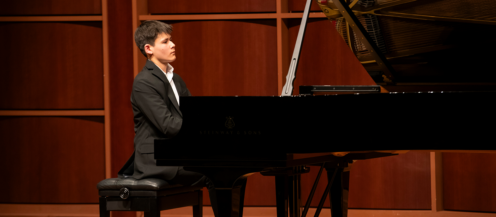 Young pianist seated at a Steinway & Sons grand piano, performing on a concert hall stage.