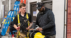 Two men work together in a workshop; one wears safety gear and the other adjusts equipment on a yellow machine.