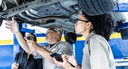 Instructor and students examining the underside of a car on a lift in an automotive training workshop.