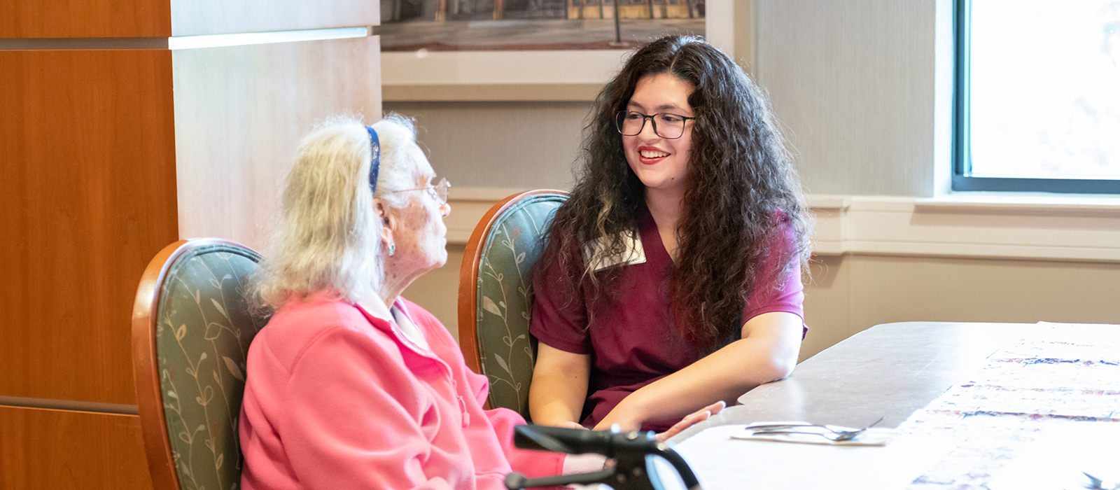 Young caregiver in scrubs smiling and talking with an older woman in a senior care setting, seated together at a table in a warmly lit room.