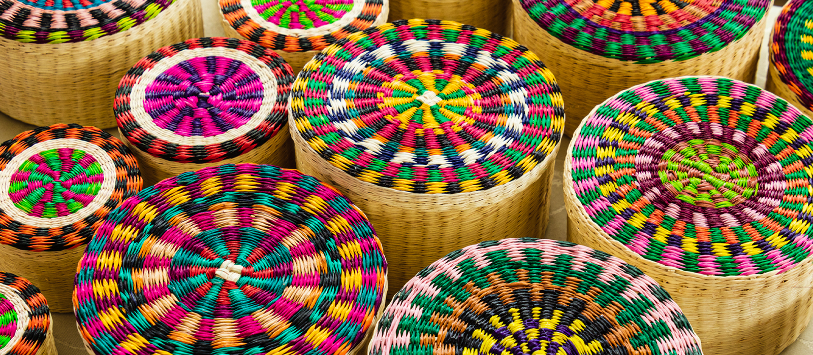 A collection of handcrafted woven baskets with intricate, colorful patterns on their lids. The baskets are made from natural fibers and feature bright hues of pink, green, blue, yellow, orange, and black, creating eye-catching geometric designs.