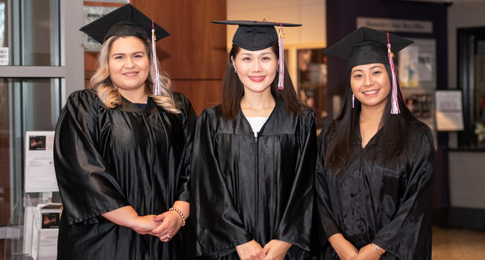 Three graduates in caps and gowns smiling together in a campus lobby