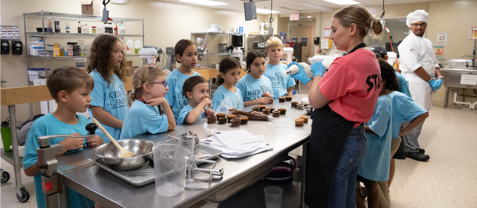 An adult instructor wearing a coral shirt teaching a baking class full of young students wearing light blue shirts in a culinary kitchen.