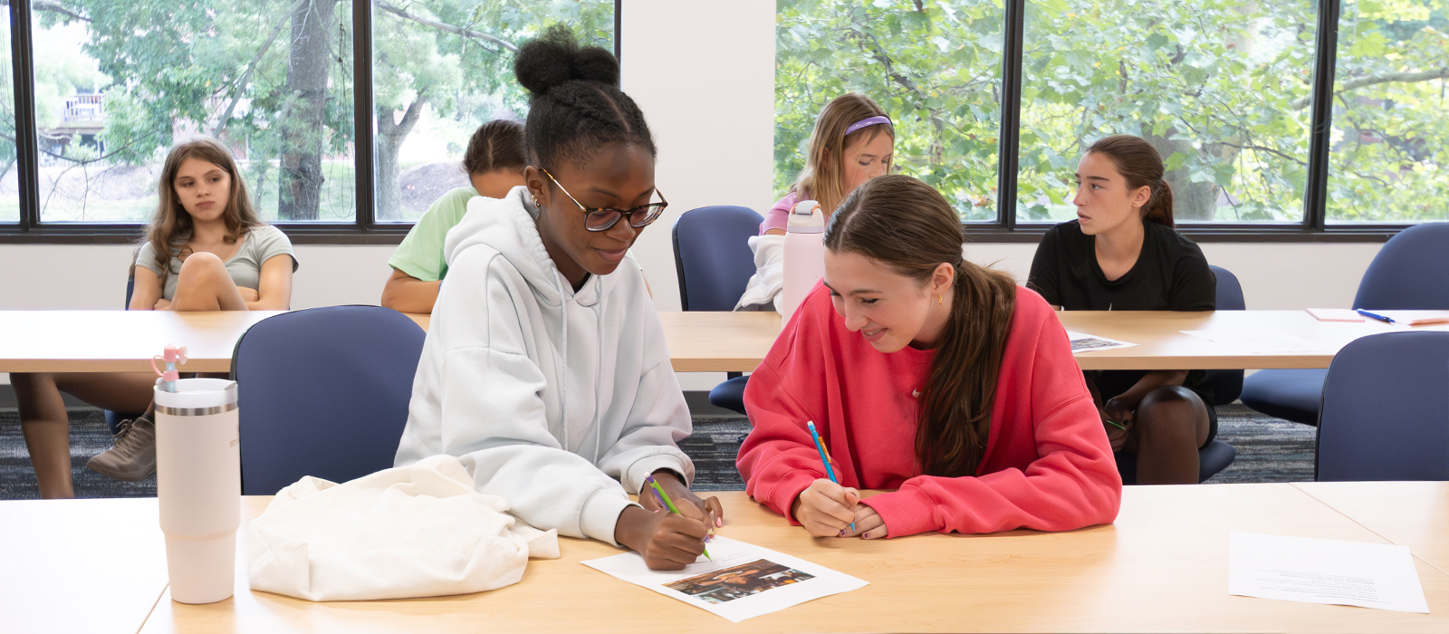 Students sitting in a bright classroom collaborate at a table, smiling as they work together on an assignment, with others engaged in activities in the background.