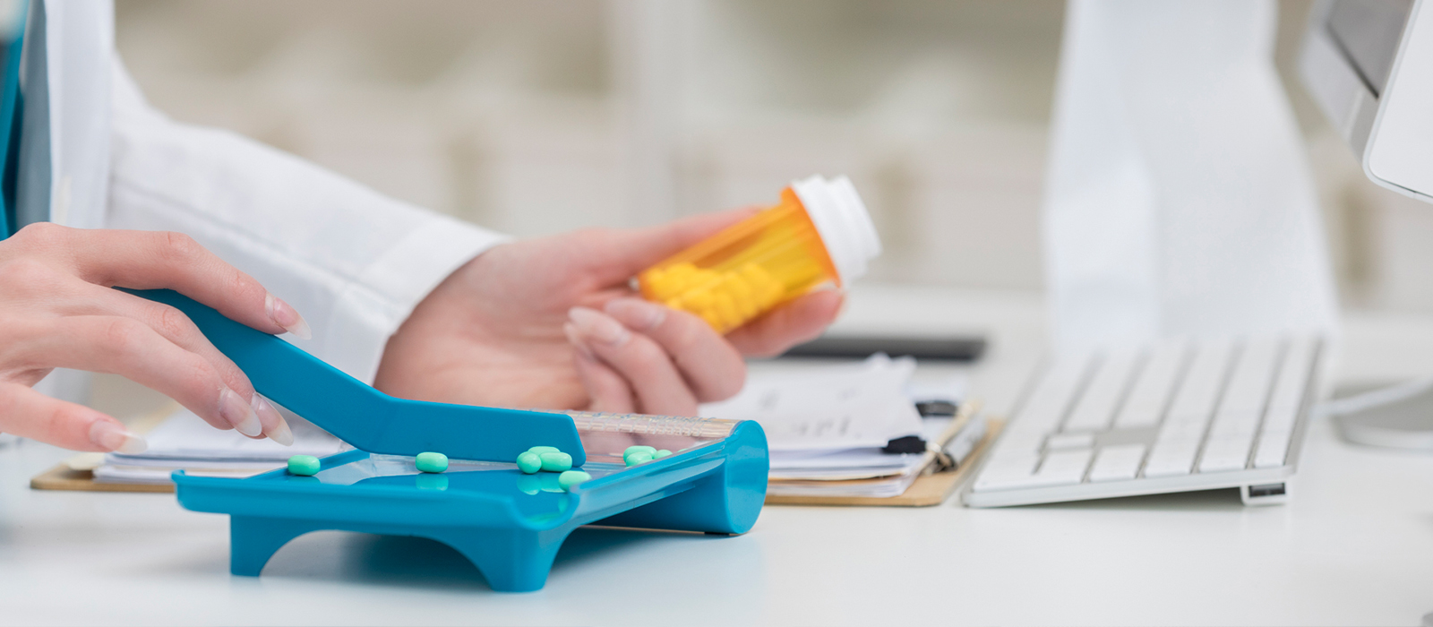 A healthcare trainee practices counting prescription pills on a pharmacy tray while holding a medication bottle next to a computer in a clinical training setting.