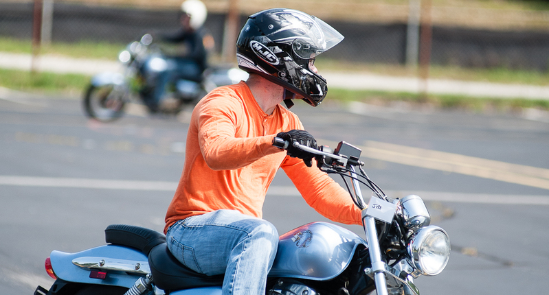 Person wearing a black full-face motorcycle helmet and orange shirt sitting on a light-blue cruiser motorcycle in a parking lot