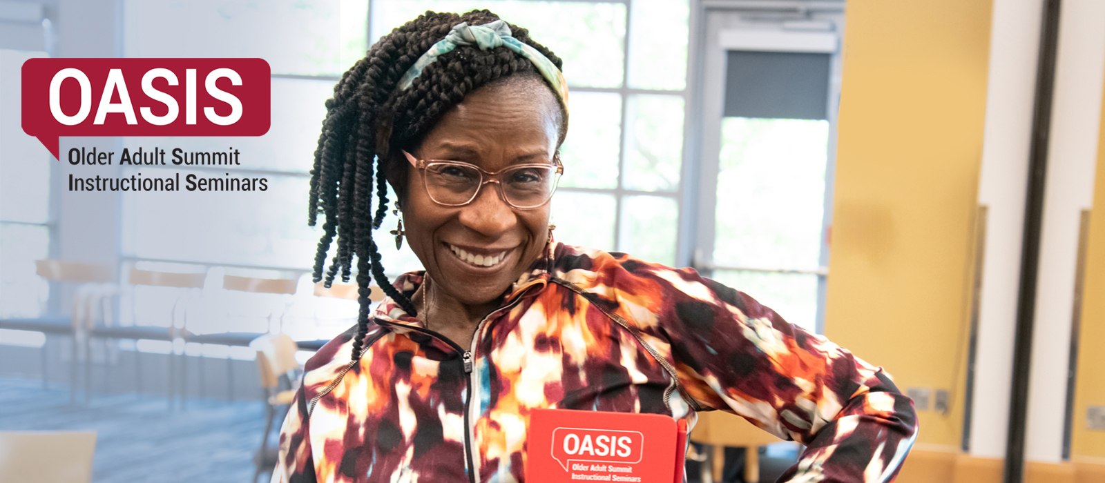 Smiling woman wearing glasses and a colorful jacket holds an OASIS sign at the Older Adult Summit Instructional Seminars event.