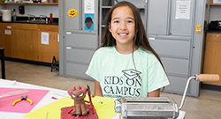 A young student wearing a mint green 'Kids on Campus' shirt in a classroom pottery class.
