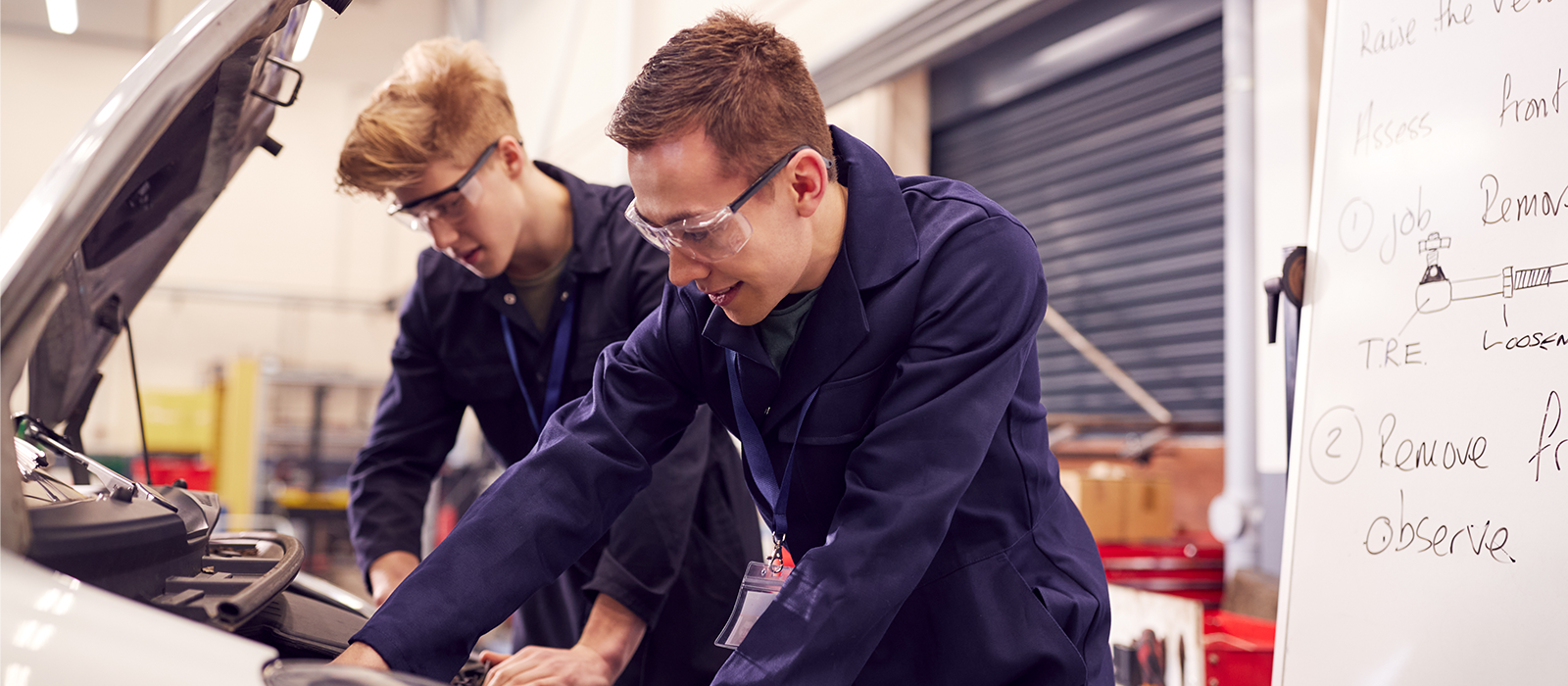 Two men engaged in car repairs inside a garage, with various tools and car parts visible in the background.