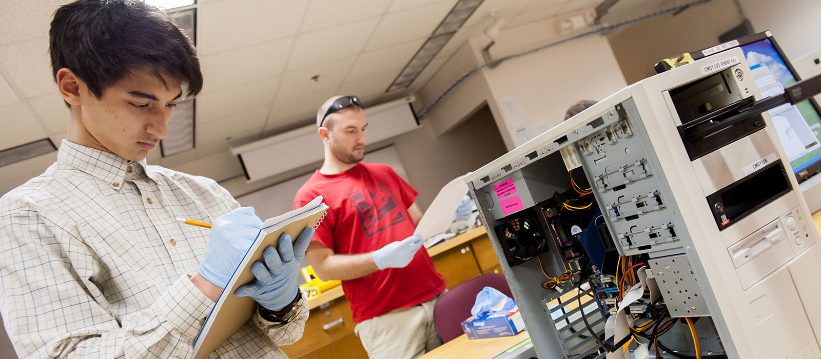 Two student wearing rubber gloves standing over computer equipment
