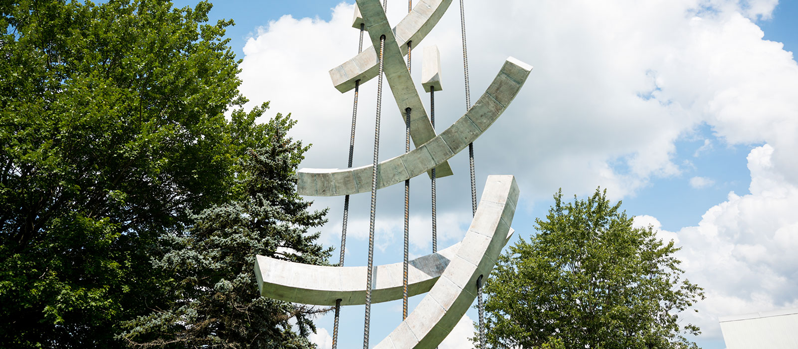 A modern metal sculpture in front of trees and partly cloudy skies