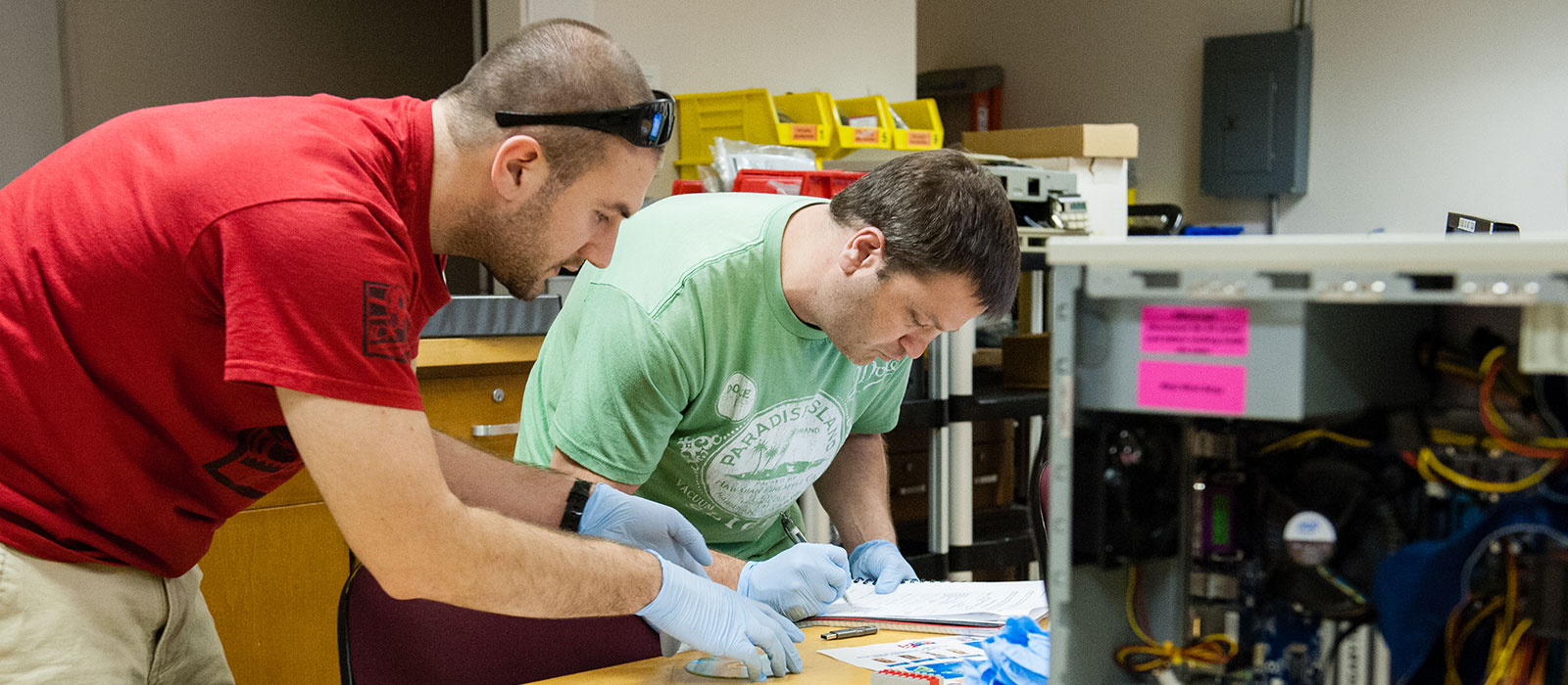 Two students wearing rubber gloves standing over computer equipment