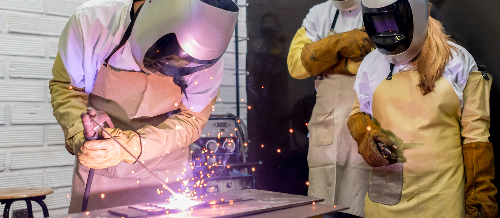 Welders focused on a metal object, with bright sparks flying as they perform welding tasks in a busy workshop.