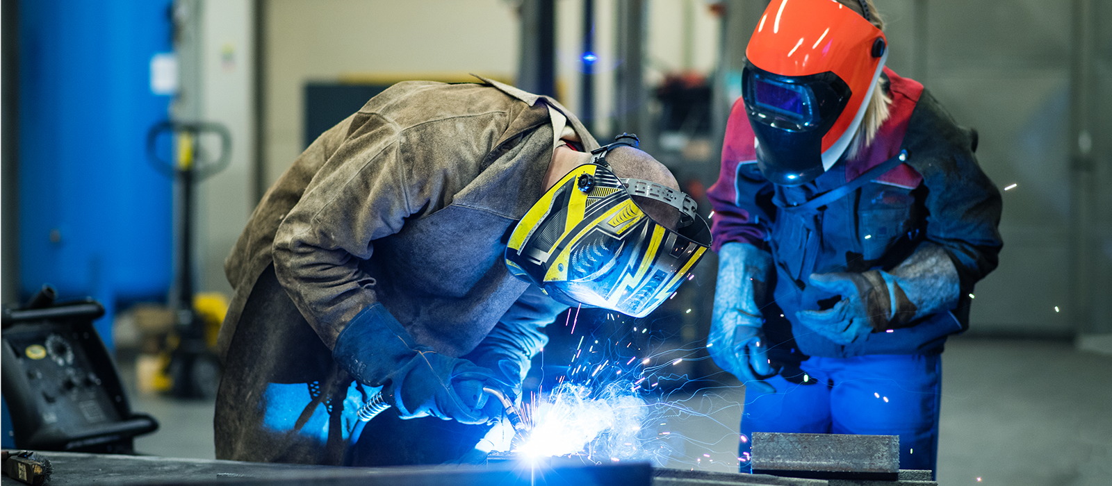 Two men wearing safety gear perform welding on metal pieces inside a factory environment.