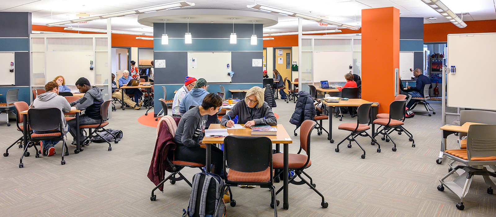 A modern educational space with students and instructors working collaboratively at tables. The room has an open layout with whiteboards, large windows, and bright orange and blue accent walls. Groups of students are engaged in discussions, writing, and using laptops, while others are seated individually. The space is well-lit, with pendant lights and natural lighting.