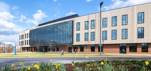 Exterior view of the Kahlert Foundation Complex on a sunny day, featuring a modern three-story building with large glass windows. Yellow daffodils bloom in the foreground, and a few people are walking along the sidewalk.
