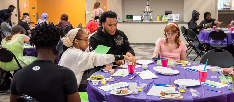Students sit around a table covered with art supplies, engaging in a creative activity during a campus event.
