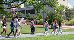 A group of students walks together across a campus pathway surrounded by trees and greenery during a sunny day.