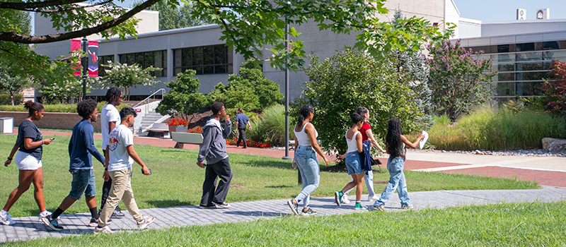 A group of students walks together across a campus pathway surrounded by trees and greenery during a sunny day.