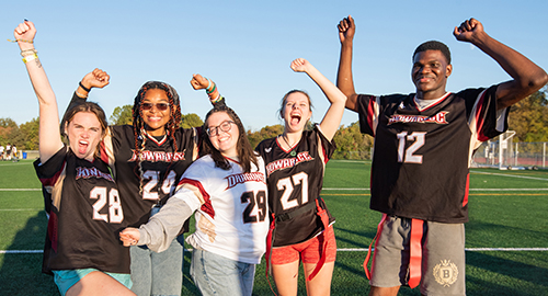 A group of five smiling students wearing sports jerseys stand together on a football field with their arms raised in celebration.